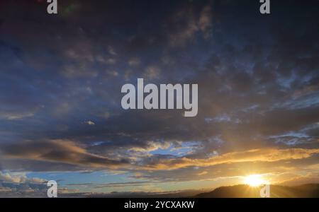 Sun setting sunburst behind mountain with blue sky and clouds tinged with colour Stock Photo