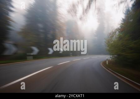 Long exposure, blurry autumn morning covered empty rural road through a forest in Europe. Wide angle view, limited visibility, bad driving weather con Stock Photo