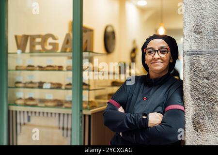 A cheerful pastry chef in a modern bakery, standing confidently with an array of vegan donuts creatively displayed in the background Stock Photo
