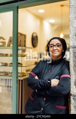 A confident vegan pastry chef stands with her arms crossed in front of her bakery boasting a variety of gourmet vegan donuts Stock Photo