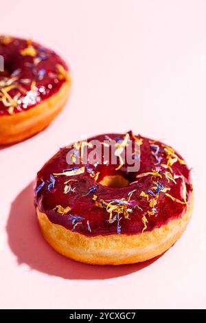 Colorful vegan donuts glazed with a rich red fruit topping and sprinkled with edible blue and yellow flowers, showcased on a light pink background Stock Photo