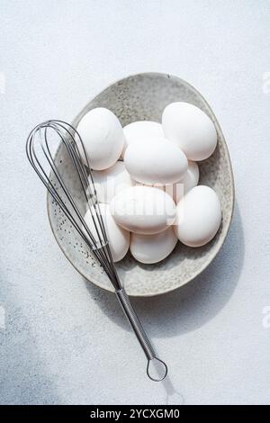 Top view of a bowl full of white fresh raw chicken eggs alongside a stainless steel whisk, set against a light textured background. Stock Photo