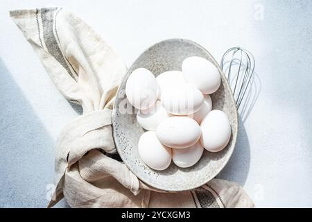 Top view of a speckled ceramic bowl filled with pristine white raw chicken eggs, accompanied by a whisk and a striped cloth, set against a bright sunl Stock Photo