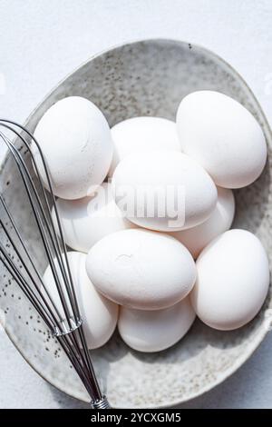 Top view of a monochromatic photo featuring a bowl full of fresh, white chicken eggs accompanied by a whisk, depicting raw ingredients in a kitchen se Stock Photo