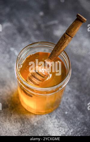 From above, organic honey drips from a wooden dipper into a glass jar, captured on a textured surface. Stock Photo