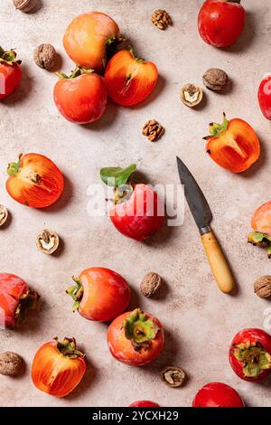 A vibrant display of whole and sliced persimmons with scattered walnuts and a rustic knife on a textured gray background, highlighting fresh, autumnal Stock Photo