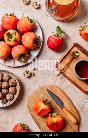 An appealing setup featuring fresh persimmons alongside walnuts and a pot of tea, captured on a warm-toned kitchen table setting Stock Photo
