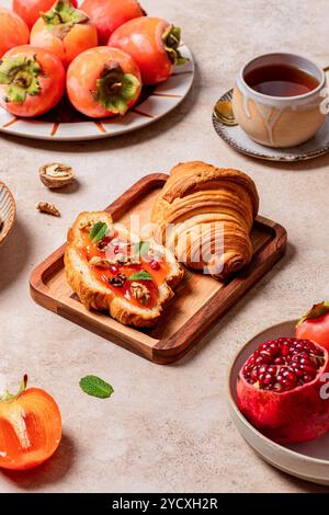 A cozy breakfast setting featuring ripe persimmons, freshly baked croissants topped with persimmon slices, walnuts, and pomegranate seeds, served alon Stock Photo