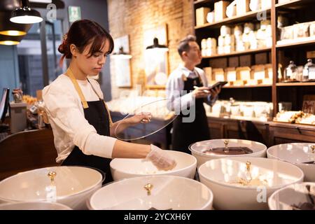 A concentrated young woman placing chocolates in large white bowls in a well-lit chocolate shop, with a male colleague using a tablet in the backgroun Stock Photo