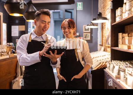 Two smiling chocolate store employees, one female and one male, collaborate using a digital tablet amidst shelves filled with chocolate products Stock Photo
