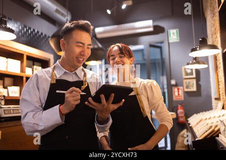 Two cheerful staff members, a man and a woman, interact over a digital tablet inside a cozy chocolate store, surrounded by shelves of assorted chocola Stock Photo