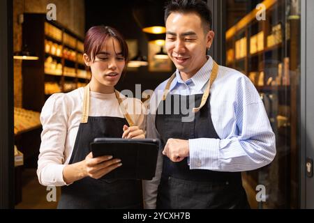 Two store clerks in a gourmet chocolate shop are reviewing inventory using a tablet Both are wearing aprons and smiling while discussing their work in Stock Photo