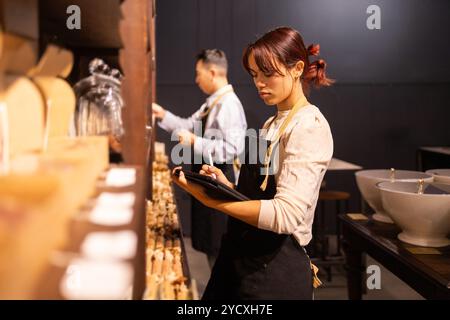 A female employee checks inventory on a tablet at a well-equipped chocolate store, while her male colleague arranges products in the background Stock Photo