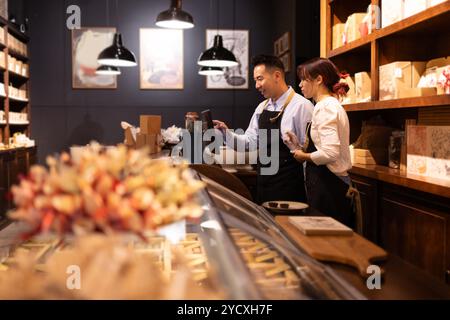 A male and female employee interact while using a computer in a cozy, well-lit chocolate shop, showcasing packaged chocolates and gifts on dark wooden Stock Photo
