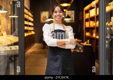 A cheerful young woman with a welcoming smile stands confidently in a cozy chocolate store, wearing a white shirt, dark apron, and crisscross suspende Stock Photo