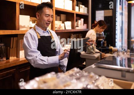 A male shopkeeper with a smiling expression operates the register at a cozy chocolate store, while a female colleague works in the background Stock Photo