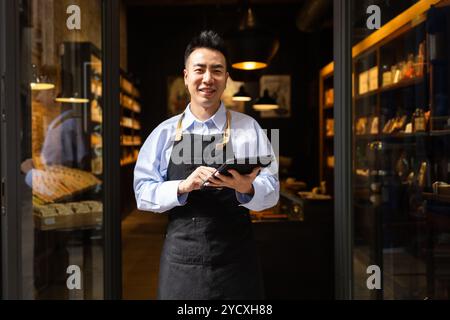 An Asian chocolate factory worker stands proudly at the entrance, holding a digital tablet Stock Photo