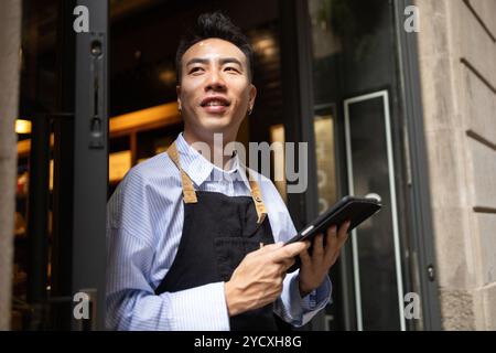 A cheerful Asian male in a striped shirt and black apron, managing a chocolate shop while using a digital tablet by the entrance Stock Photo