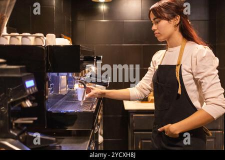 A focused female barista expertly operates an espresso machine in a dimly lit cafe, crafting a perfect cup of coffee for awaiting customers Stock Photo