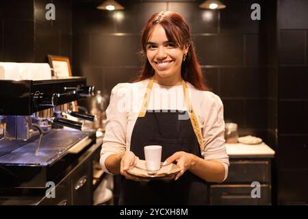 A smiling female barista in a black apron serves a cup of coffee in a chic, dark-toned chocolate store Stock Photo