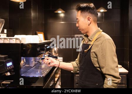 Asian Barista in apron skillfully operates a coffee machine inside a modern, dark-themed cafe, showcasing his expertise in coffee preparation Stock Photo