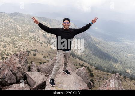 Joyful male hiker raising arms in victory atop a jagged mountain peak, overlooking the lush, forested valleys of Cumbres del Ajusco, national park in Stock Photo