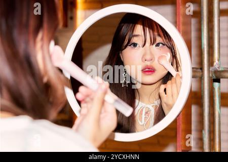 A Chinese Gen Z girl carefully applies makeup using a brush, as seen in the reflective surface of a round mirror, highlighting a moment of beauty rout Stock Photo