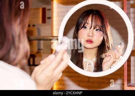 A young Chinese Gen Z girl carefully applies makeup while looking into a round mirror, surrounded by beauty products in a cozy indoor setting Stock Photo