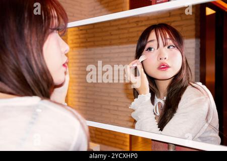 A young Chinese Gen Z girl carefully applies makeup while looking into a mirror, capturing a moment of everyday beauty and self-expression Stock Photo