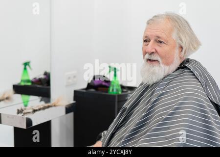 An elderly man with a content expression sits in a barbershop, having donated his hair for a cause Cut locks of hair are visible on a nearby glass tab Stock Photo