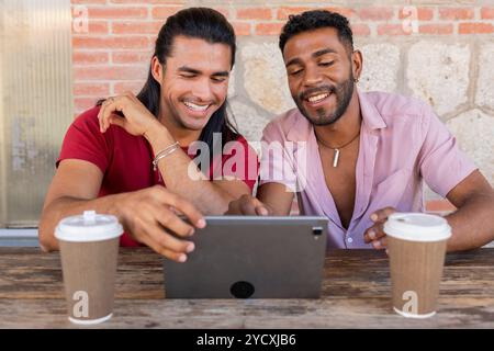 Gay couple, one Hispanic and the other African American, sharing a light moment over coffee while using a digital tablet outdoors in an urban setting, Stock Photo