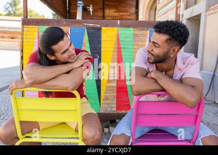 A joyful gay couple, one Latin American and the other African American, share a tender moment seated on vibrant chairs in an urban environment, symbol Stock Photo