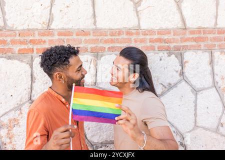 Two joyous men, reflecting a multiethnic and gay American Latin identity, share a light-hearted moment while holding a pride flag against an urban bri Stock Photo