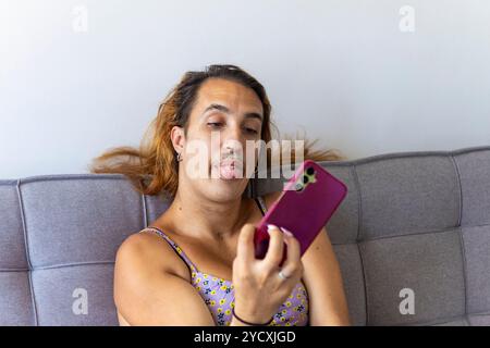 A trans woman in a floral top, on a gray sofa, sticking out her tongue showing her piercing while looking at her smartphone This image captures a rela Stock Photo