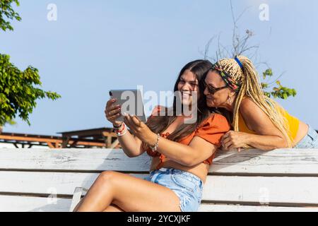 A loving mother and her transgender daughter, formerly her son, are happily using a digital tablet while sitting outdoors on a bench, sharing a joyful Stock Photo