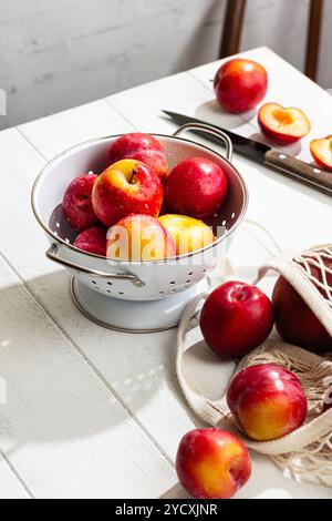 Juicy red plums, freshly washed with visible water droplets, are stacked in a white colander on a light wooden table. Stock Photo
