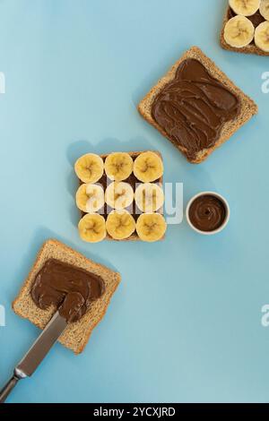 Overhead view of a simple, nutritious breakfast with whole-grain bread, chocolate spread, and sliced bananas on a pale blue background Stock Photo
