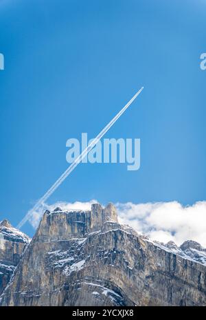 An airplane leaves a white contrail above the rugged peaks of the Swiss and French Alps under a clear blue sky. Stock Photo