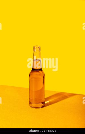 A clear glass bottle with a beer sits on a textured surface against a vivid yellow backdrop, creating a minimalistic yet striking visual contrast Stock Photo