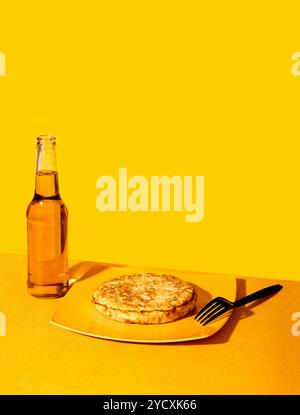 A monochromatic image featuring a glass bottle of beer and a Spanish-style tortilla on a bright yellow plate, all against a yellow backdrop, highlight Stock Photo