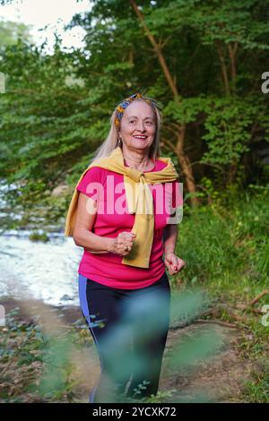 Senior woman smiling while jogging near a stream in a forest park, dressed in a vibrant sporty outfit and adorned with a colorful headband Stock Photo