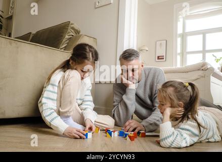 A father and his two daughters are playing a game with colorful wooden blocks on the floor. The father is lying on the floor, watching as his daughter Stock Photo