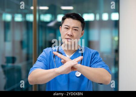 Asian doctor in blue scrubs highlights nonverbal communication in healthcare by crossing hands, demonstrating seriousness and professionalism in clinical setting, using effective body language. Stock Photo