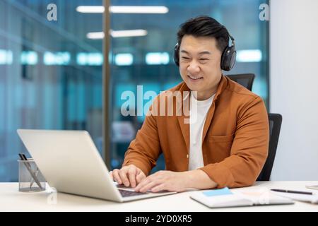 Asian businessman with headphones working on laptop showcases focus, productivity in office. Casual attire signifies modern work culture. Conceptualizes technology-driven meetings, coding work. Stock Photo