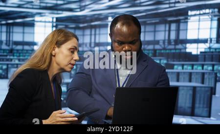 Teamworking technicians managing data center with advanced technology using devices. Coworkers in server room optimizing data center infrastructure with laptop and tablet, camera A Stock Photo