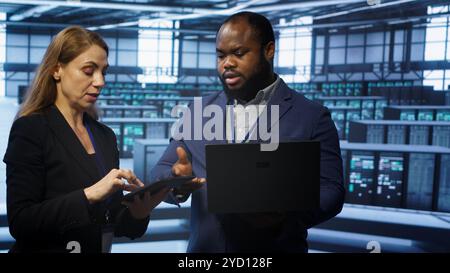 Teamworking colleagues doing checkup, inputting data on laptop of needed server hub fixes. Coworkers working together in data center housing advanced storage infrastructure, camera A Stock Photo