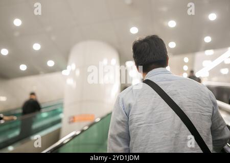 Man wearing a blue shirt and a black backpack is walking down an escalator. There are other people in the background Stock Photo