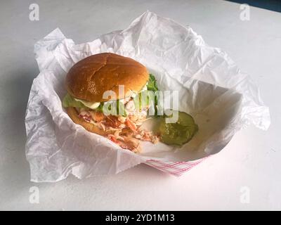 A lobster roll, dripping with mayonnaise and with a sliced pickle in a basket. At Young's Lobster Pound at the harbor of Belfast, Maine, USA. Stock Photo