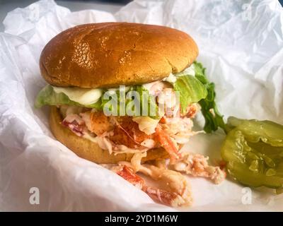 A lobster roll, dripping with mayonnaise and with a sliced pickle in a basket. At Young's Lobster Pound at the harbor of Belfast, Maine, USA. Stock Photo