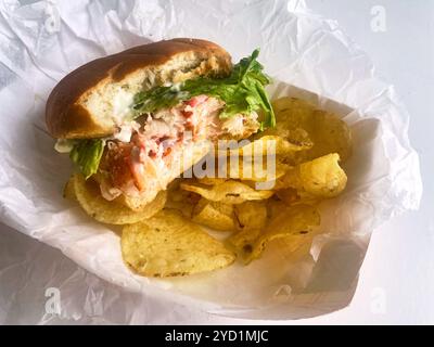 A lobster roll with a bite out of it with kettle potato chips in a basket. At Young's Lobster Pound at the harbor of Belfast, Maine, USA. Stock Photo